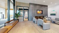 Located at 325 Royal Commons in Victoria, BC, this contemporary building lobby offers a refined and welcoming arrival experience. The space is defined by soaring double-height ceilings and a sophisticated palette of materials, including large-format concrete-look floor tiles, smooth rendered walls, and warm wood slat ceiling panels that add texture and contrast throughout.

The main lounge area centers around a sleek, wide-format electric fireplace set within a floor-to-ceiling dark panel feature wall, accompanied by a mounted display screen and a curated arrangement of soft, curved upholstered armchairs over a neutral area rug. Potted tropical plants are positioned throughout, adding life and a natural element to the modern interior.

An entrance corridor connects to the building's glass double doors, framed by large floor-to-ceiling windows that invite abundant natural light and provide views of the surrounding streetscape. A built-in wooden bench offers a practical seating option near the entry.

The service zone includes a bank of smart parcel lockers for secure package management alongside a stainless steel elevator, all set beneath the dramatic volume of the double-height ceiling. An additional glassed-in room with vertical slat detailing and circular wall sconces is accessible from the lounge, providing further amenity space. Together, these elements create a polished and functional lobby befitting a modern residential building.