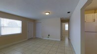 This single-story home presents a straightforward and functional layout finished with consistent light beige ceramic tile flooring throughout the interior. The main living area is open and bright, painted in neutral, warm tones that lend a clean and welcoming ambiance. A flush-mount ceiling light fixture illuminates the space, complemented by a textured ceiling finish. Practical touches include a standard closet with louvered bi-fold doors, providing accessible storage, alongside a solid panel interior door. Utility connections are visible along the baseboard, indicating laundry or appliance hookup access within the living space. A central hallway connects the main area to additional rooms, with natural light visible at the far end through a window, suggesting well-distributed illumination across the floor plan. White baseboards trim the walls throughout, adding a crisp contrast to the warm wall color. HVAC vents are integrated into both the ceiling and walls, ensuring climate control throughout. The overall design is simple, clean, and move-in ready, suited for a variety of uses and configurations.
