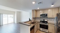 This single-floor space showcases a thoughtfully designed open-concept layout that seamlessly connects the kitchen and living areas, creating a bright and cohesive environment. The contemporary kitchen is a standout feature, equipped with stainless steel appliances including a gas range, built-in microwave, and dishwasher. Light wood cabinetry provides ample storage while lending warmth to the space, complemented by rich dark stone countertops that extend across the kitchen peninsula. A mosaic tile backsplash in neutral tones adds a refined textural detail, tying the design elements together. The peninsula doubles as a breakfast bar, enhancing the kitchen's functionality and its connection to the open living area beyond. Recessed lighting illuminates the space efficiently, while light-toned hardwood flooring runs throughout, contributing to the airy and modern aesthetic. The living area benefits from natural light filtering in through what appears to be a sliding door or large window, adding brightness and a sense of openness to the overall floor plan. Clean white walls and a neutral palette throughout make the space feel fresh, versatile, and move-in ready.