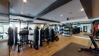 Located at 330 Westlake Ave in Los Angeles, CA, this fitness center is a thoughtfully designed, single-floor facility that offers a comprehensive workout experience. The space is defined by warm hardwood-style flooring, crisp white walls, and striking dark exposed ceiling beams that lend an industrial-modern aesthetic throughout.

The main workout floor is generously sized, accommodating a robust selection of strength and cardio equipment. A row of treadmills and elliptical machines lines one side, while a dedicated free weights area features a full dumbbell rack, a stair-climber trainer, and a power rack for heavy lifting. Cable and multi-station machines provide additional strength training options, and floor-to-ceiling mirrors along one wall visually expand the space while allowing for form checks during exercise.

Large sliding glass windows along the perimeter flood the cardio zone with natural light and provide views of the surrounding neighborhood, creating an open and energizing atmosphere. A separate yoga and stretching studio offers a quieter environment, furnished with TRX mats and a wall-mounted flat-screen TV, with its own large windows bringing in ample daylight.

The entry area includes a built-in cubby console, a wall-mounted TV, a built-in water station, and decorative greenery, creating a welcoming transition into the facility. Recessed lighting and ceiling fans complete the polished, functional environment.