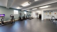 Located at 2020 4th St in Berkeley, CA, this fitness center offers a well-appointed exercise space designed to support a comprehensive workout experience. The single-floor layout is thoughtfully organized to accommodate a wide variety of training styles, providing ample room to move freely between equipment zones.

The cardio area features multiple treadmills and elliptical machines, each equipped with individual display screens, positioned along a teal-painted wall with windows that allow natural light to filter into the space. Overhead, bright fluorescent lighting ensures consistent illumination throughout.

The strength training area is equally well-equipped, housing a multi-station cable machine, selectorized weight machines, and a dedicated free weights section complete with a dumbbell rack and flat benches. Full-length mirrors line the walls in this zone, enhancing the sense of openness while allowing users to monitor their form during exercise.

Durable dark rubber flooring covers the entire space, providing both safety and comfort underfoot. Structural columns are cleanly finished in white, contributing to the clean, modern aesthetic of the room. The walls feature a neutral white and muted teal color scheme that gives the space a calm, focused atmosphere. A wall-mounted display and decorative artwork add subtle character to the environment, making this fitness center both functional and inviting.