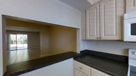 This two-floor residence offers a practical and well-organized layout designed for comfortable everyday living. The kitchen is a standout space, appointed with crisp white raised-panel cabinetry that provides generous storage both above and below the counters. Deep, dark granite countertops contrast cleanly with the light cabinetry, lending the kitchen a polished and durable finish. A defining architectural feature of the kitchen is the open pass-through cutout in the wall, which connects directly to the adjacent living area. This thoughtful design element promotes an open, communicative flow between the cooking and living spaces, allowing natural sightlines and easy interaction between rooms. The living area, visible through the pass-through, features neutral-toned walls and tiled flooring, contributing to a clean and understated aesthetic throughout. The two-floor layout of the residence provides a natural separation between living and private spaces, offering both functionality and versatility. Overall, the space presents a straightforward and well-maintained interior with classic design choices, including neutral color palettes and timeless finishes, making it adaptable to a variety of furnishing styles and personal tastes.