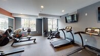 This fitness center at 700 Emerald St SE in Saint Paul, Minnesota, is a thoughtfully designed, single-floor facility that provides a comprehensive workout environment. The space features a clean, modern aesthetic with cool gray and bold terracotta-orange accent walls, recessed lighting, and durable rubber-composite flooring suited for high-traffic exercise use.

The cardio zone is well-appointed with multiple treadmills, stationary bikes, and rowing machines, all positioned to take advantage of large windows that invite natural light and views of the surrounding outdoor area. Wall-mounted flat-screen televisions are strategically placed throughout the space, allowing users to stay entertained during workouts.

The strength training area is stocked with a full array of Life Fitness selectorized machines, free weights including a color-coded dumbbell rack, and workout benches, catering to a wide range of fitness goals. Full-length mirrors line one wall, enhancing the sense of openness while allowing users to monitor their form.

Tasteful framed artwork adds a welcoming touch to the walls, contributing to a polished, motivating atmosphere. The layout is open and well-organized, ensuring ease of movement between equipment zones. With ample natural light, modern finishes, and a diverse selection of equipment, this fitness center offers a functional and inviting space for a complete workout experience.