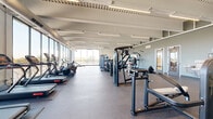 Located at 2800 N Wayzata Blvd in Minneapolis, MN, this expansive fitness center spans a single floor and offers a thoughtfully designed exercise environment suited for a wide range of training needs. The space is defined by its modern architectural character, featuring high exposed ceilings with structural beams, linear LED lighting, and large-scale floor-to-ceiling windows that flood the interior with natural light and provide sweeping outdoor views of the surrounding landscape.

The main exercise floor is generously proportioned, accommodating a robust lineup of strength training machines, including cable systems, leg press equipment, and multi-function units, as well as a dedicated free weights area stocked with dumbbells, medicine balls, and a flat bench. A mirrored wall in the free weights zone enhances the sense of space and supports proper form during workouts.

A well-equipped cardio zone lines the window wall, featuring treadmills, ellipticals, stationary bikes, and rowing machines — all positioned to take advantage of the natural light and outdoor views. The rubber flooring throughout provides both durability and comfort underfoot.

A separate area near the entrance features a smooth epoxy-style floor, built-in storage cubbies, and a water station, serving as a transition and amenity zone. Glass partition walls and doors connect distinct areas of the facility while maintaining an open, airy atmosphere throughout.