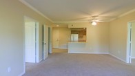 This single-floor space features an open-concept layout that seamlessly connects the living area to the kitchen, creating a cohesive and functional floor plan. The living area is finished with wall-to-wall carpeting in a neutral beige tone, contributing to a warm and understated ambiance. Crown molding runs along the ceiling perimeter, adding a subtle architectural detail, while a ceiling fan with a light fixture is centrally positioned to provide both comfort and illumination.

The kitchen is situated at the rear of the main living space and is defined by a raised breakfast bar-style peninsula, which serves as a natural dividing element between the two areas. Inside the kitchen, wood-toned cabinetry lines the upper walls, accompanied by a full-size refrigerator and what appears to be a standard range, along with track lighting overhead. Recessed lighting further brightens the kitchen zone.

A hallway visible on the left side of the space leads to additional rooms, with multiple white panel doors suggesting access to bedrooms or other private areas. The overall color palette consists of soft, neutral tones on the walls and trim, lending the space a clean and move-in-ready appearance.