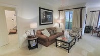 This single-floor living space showcases a carefully designed living room that balances comfort and style. The room features crown molding along the ceiling, lending a refined architectural detail to the overall aesthetic. Neutral walls in a soft, warm tone provide a versatile backdrop that complements the rich brown upholstered sofa, which serves as the room's centerpiece and is accented with decorative striped and solid throw pillows.

Natural light filters through a window dressed with cream drapery panels and horizontal blinds, offering a view of lush greenery outside. A patterned accent chair positioned near the window adds visual interest and texture to the space. The glass-topped coffee table with a dark metal frame provides both functionality and an open, airy feel to the seating area.

Ambient lighting is provided by a classic table lamp on a small round side table, which also holds a decorative floral arrangement. A large framed black-and-white photograph serves as a focal wall accent above the sofa, adding a sophisticated artistic element. Light-toned carpeting runs throughout, contributing to the overall warmth and comfort of the space.