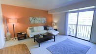 This single-floor residence presents a cohesive and thoughtfully styled interior defined by warm terracotta-orange accent walls that create a welcoming atmosphere throughout. The living area is carpeted and comfortably proportioned, anchored by a sectional sofa and a dark wood coffee table. A large sliding glass door floods the room with natural light and opens directly onto a private balcony, offering outdoor views and an airy feel. A decorative triptych floral canvas above the sofa adds an artistic touch, while a patterned area rug and coordinated furnishings contribute to the polished presentation.

The kitchen and dining area share an open layout with contrasting dark and white cabinetry and a peninsula counter that separates the cooking space from the dining zone. A compact white dining table with drop-leaf functionality maximizes the use of space, accompanied by dark cross-back chairs. Open wall shelves display dishware and decorative vases, and a dark console table serves as a functional sideboard. Hardwood-style flooring runs through the kitchen and dining area, while a ceiling fan provides additional comfort. The overall ambiance is warm, modern, and efficiently organized.