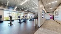 Located at 710 Vikings Pkwy in Eagan, Minnesota, this fitness facility offers a generously sized, single-floor layout designed to accommodate a full spectrum of workout needs. The open-concept floor plan is anchored by an expansive main workout area featuring high, exposed truss ceilings with industrial steel framework and bright linear lighting that creates a clean, energizing atmosphere throughout.

Large floor-to-ceiling windows line the exterior walls, flooding the space with abundant natural light and offering views of the surrounding greenery — enhancing the overall workout environment. Durable rubber flooring throughout the main areas provides both functionality and safety for high-impact activities.

The cardio zone is well-appointed with rows of treadmills, stationary bikes, and elliptical machines, each equipped with individual display screens. A dedicated strength training area features a comprehensive selection of cable machines, free weights, dumbbell racks, and power racks, all set against bold purple accent walls with wall-mounted televisions. An additional open functional training area offers power racks, benches, and a full dumbbell collection for versatile workouts.

A separate group fitness studio features smooth wood-look flooring, full-wall mirrors, and a drop ceiling — providing a quieter, more intimate setting for classes and personal training sessions, complete with stability balls and various small equipment storage.