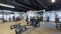 This fitness facility offers a comprehensive workout environment designed to accommodate a wide range of exercise needs. The space features an open, industrial-style aesthetic, with exposed black-painted ductwork and structural elements overhead contrasting against clean white columns and walls, creating a modern and functional atmosphere. The flooring consists of durable rubber tiles, providing safety and support throughout the workout area. Large wall-mounted mirrors are strategically placed to allow members to monitor their form during exercises and to enhance the sense of space throughout the facility. The gym is outfitted with an extensive selection of professional-grade strength training machines, including cable machines, chest press, and leg equipment, as well as adjustable benches suitable for a variety of free weight exercises. Overhead industrial pendant lighting ensures the space is well-lit throughout. The layout is thoughtfully organized, with equipment arranged to allow for comfortable movement and accessibility between stations. Additional cardio machines are visible in the background, rounding out the facility's offerings. Overall, the space delivers a serious, performance-focused training environment suitable for users of all fitness levels.