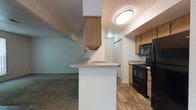 This single-floor unit offers a thoughtfully laid-out interior that balances functionality with comfort. The entryway greets visitors with tile flooring that transitions seamlessly into carpeted living areas, establishing a clear sense of arrival and flow throughout the space.

At the heart of the unit is an open-concept kitchen and living area. The kitchen is well-equipped with black appliances, including a gas range, over-the-range microwave, and refrigerator, complemented by warm wood-toned cabinetry. A peninsula with a light-toned countertop serves as both a workspace and a visual divider between the kitchen and the adjacent living area. An overhead cabinet suspended above the peninsula adds additional storage while maintaining the open feel of the space. The kitchen features wood-style flooring, providing a durable and clean aesthetic.

The living area flows naturally from the kitchen, offering a comfortable, carpeted expanse with a neutral color palette and clean white walls. A large sliding glass door or wide window with vertical blinds invites abundant natural light and outdoor views.

The bedrooms are carpeted and well-proportioned, each featuring windows that frame greenery views outside, filling the rooms with soft, natural light. The overall ambiance of the unit is clean, practical, and move-in ready, with a neutral design that offers a versatile backdrop for personalization.