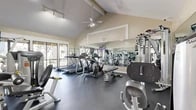 Located at 401 W Orangewood Ave in Anaheim, California, this fitness center offers a well-equipped exercise space designed to support a comprehensive workout routine. The room is defined by its striking vaulted ceiling with exposed beams, giving the space a sense of openness and volume well beyond its footprint. A ceiling fan circulates air throughout, contributing to a comfortable environment during workouts.

Natural light streams in through large sliding glass doors and windows along one wall, providing views of the surrounding outdoor greenery and keeping the atmosphere bright and inviting. Track lighting supplements the natural light, ensuring even illumination across the entire floor.

The exercise room is outfitted with a well-rounded selection of Hoist-brand strength training machines, including cable systems and seated resistance equipment, alongside multiple treadmills, an elliptical trainer, and a stationary bike. Full-length mirrored walls on two sides visually expand the space and allow users to monitor their form during exercise. The durable dark rubber flooring provides cushioning and stability underfoot, suitable for both cardio and strength activities.

Wall-mounted televisions add an entertainment element for those engaged in longer cardio sessions. The overall ambiance is clean, functional, and professional, reflecting a thoughtfully designed fitness facility.