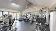Located at 401 W Orangewood Ave in Anaheim, California, this fitness center offers a well-equipped exercise space designed to support a comprehensive workout routine. The room is defined by its striking vaulted ceiling with exposed beams, giving the space a sense of openness and volume well beyond its footprint. A ceiling fan circulates air throughout, contributing to a comfortable environment during workouts.

Natural light streams in through large sliding glass doors and windows along one wall, providing views of the surrounding outdoor greenery and keeping the atmosphere bright and inviting. Track lighting supplements the natural light, ensuring even illumination across the entire floor.

The exercise room is outfitted with a well-rounded selection of Hoist-brand strength training machines, including cable systems and seated resistance equipment, alongside multiple treadmills, an elliptical trainer, and a stationary bike. Full-length mirrored walls on two sides visually expand the space and allow users to monitor their form during exercise. The durable dark rubber flooring provides cushioning and stability underfoot, suitable for both cardio and strength activities.

Wall-mounted televisions add an entertainment element for those engaged in longer cardio sessions. The overall ambiance is clean, functional, and professional, reflecting a thoughtfully designed fitness facility.