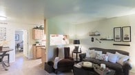 This single-floor space offers a comfortable and cohesive layout, with the living area and kitchen thoughtfully connected through an open pass-through window that promotes a sense of openness while maintaining distinct functional zones. The living area is furnished with rich, dark upholstered seating, including a tufted wingback chair and a generous leather sofa adorned with decorative pillows, creating an inviting and lived-in feel. A classic table lamp provides warm ambient lighting, while floating wall shelves display tasteful décor accents such as a vintage clock and decorative keys, adding character to the soft sage-green walls. The kitchen, visible through the pass-through, features natural wood cabinetry with a rustic, warm finish, complemented by light countertops and a tile backsplash. Upper cabinets extend to the ceiling, maximizing storage. Potted greenery is incorporated throughout, bringing subtle life and freshness to the interior. The light-toned flooring flows consistently between spaces, reinforcing the cohesive design. Overall, the space presents a cozy, rustic-inspired aesthetic with practical layout choices.