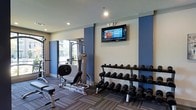 This fitness center is designed to support a comprehensive workout experience within a clean, modern environment. The space features a two-tone color scheme of cool blue accent walls paired with neutral white and gray tones, creating an energizing yet focused atmosphere. Recessed lighting throughout the ceiling ensures the room is well-lit during any hour of use.

The strength training area is well-appointed, featuring a multi-station cable machine and an adjustable workout bench, offering a variety of resistance exercises. A full tiered dumbbell rack lines one wall, stocked with a wide range of weights to accommodate users of all fitness levels. A wall-mounted flat-screen television is positioned prominently for entertainment during workouts.

The flooring consists of durable, patterned carpet tiles suited for a fitness environment, providing both comfort and traction underfoot. A window along one wall allows natural light to filter into the space, offering views to the exterior and enhancing the overall ambiance. The layout is thoughtfully organized to maximize usability while maintaining open circulation throughout the floor, making it a functional and inviting facility for daily exercise routines.