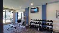 This fitness center is designed to support a comprehensive workout experience within a clean, modern environment. The space features a two-tone color scheme of cool blue accent walls paired with neutral white and gray tones, creating an energizing yet focused atmosphere. Recessed lighting throughout the ceiling ensures the room is well-lit during any hour of use.

The strength training area is well-appointed, featuring a multi-station cable machine and an adjustable workout bench, offering a variety of resistance exercises. A full tiered dumbbell rack lines one wall, stocked with a wide range of weights to accommodate users of all fitness levels. A wall-mounted flat-screen television is positioned prominently for entertainment during workouts.

The flooring consists of durable, patterned carpet tiles suited for a fitness environment, providing both comfort and traction underfoot. A window along one wall allows natural light to filter into the space, offering views to the exterior and enhancing the overall ambiance. The layout is thoughtfully organized to maximize usability while maintaining open circulation throughout the floor, making it a functional and inviting facility for daily exercise routines.