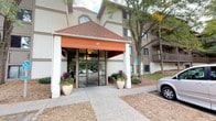Located within a multi-story building marked by a distinctive covered entryway with a terracotta-hued hip roof and an orange fabric awning, this single-floor residence offers a well-organized and functional layout. Large decorative planters flank the glass entrance doors, contributing to the building's welcoming curb appeal, which is further enhanced by mature trees and maintained landscaping.

Inside, the hallway connects the living areas with a practical flow, featuring neutral carpet flooring, crisp white walls, and dark wood door trim throughout. A set of louvered bi-fold closet doors in the hallway provides convenient storage access. The bedroom is a comfortable, softly lit space with plush carpeting, a textured ceiling, and a generously sized louvered closet. A large window fitted with vertical blinds allows natural light to enter, while a wall-mounted air conditioning unit and baseboard heating ensure year-round climate comfort. The warm, neutral tones of the walls create a calm ambiance throughout. Decorative touches, including an ornate metalwork wall medallion, add character to the interior and reflect a thoughtfully curated aesthetic.