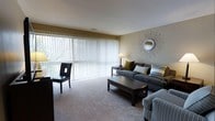 This single-floor space offers a thoughtfully designed living area that balances comfort and style. The living room serves as the centerpiece of the space, highlighted by a expansive floor-to-ceiling vertical blind-covered sliding glass door that bathes the room in abundant natural light while providing views of the surrounding greenery outdoors. The neutral-toned carpet flooring adds warmth and continuity throughout the room, complementing the soft beige walls. Seating is arranged around a dark wood coffee table, with a plush gray sofa accented by coordinating throw pillows and a striped upholstered chair, creating an inviting and cohesive conversational setting. A sleek black accent chair adds a touch of contrast near the window. Decorative touches, including a sunburst mirror mounted on the wall and a classic table lamp on a side table, contribute to the refined yet approachable ambiance of the space. The clean lines and understated color palette give the room a timeless, well-maintained aesthetic. Overall, the space presents a comfortable and functional single-level layout with an emphasis on natural light and tasteful interior design.
