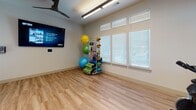 This single-floor fitness studio is thoughtfully designed to support a variety of workout routines in a clean, energizing environment. The space is divided into two distinct functional zones, connected by a bold orange-framed glass partition that adds a striking pop of color while maintaining an open, transparent feel throughout.

The main exercise room features light wood-look flooring, white walls, and an abundance of natural light streaming through multiple large windows fitted with white plantation shutters. Ceiling fans keep the space comfortable during active sessions, while overhead linear lighting ensures excellent visibility. A wall-mounted flat-screen display provides on-demand workout guidance, and a well-stocked equipment rack holds an assortment of medicine balls, resistance bands, stability balls, and exercise mats — offering everything needed for functional and cardio training.

A separate, more intimate area houses stationary cycling bikes positioned beneath their own ceiling fan, with an interior window overlooking the main exercise floor. This clever design allows for a sense of connection between zones without sacrificing dedicated focus areas.

Entry to the studio is through full-height glass doors framed in vivid orange, reinforcing the space's modern, active aesthetic. The overall ambiance is bright, motivating, and professionally equipped — ideal for group fitness classes or individual training sessions.