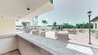 This single-level outdoor amenity space is designed for communal gathering and entertaining in a tropical setting. The covered interior area features warm terracotta tile flooring, stucco walls painted in soft coral and yellow tones, and a built-in counter with a utility sink — ideal for food and beverage service. The space opens seamlessly to a wraparound covered bar counter finished in light marble-style tile, providing ample surface space for seating and socializing. Large open-air cutouts frame panoramic views of a sun-drenched courtyard patio beyond, where additional seating arrangements, planters, and a pergola structure are visible amid lush tropical landscaping. Tall palm trees punctuate the skyline, reinforcing the resort-like ambiance. The covered sections offer shade and shelter while maintaining a strong visual and physical connection to the outdoors. A set of glass-panel doors provides access to an adjacent interior space. The overall design balances functionality and leisure, making it well-suited as a poolside bar, event venue, or outdoor lounge for a residential community or hospitality property.