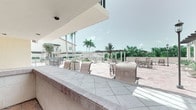 This single-level outdoor amenity space is designed for communal gathering and entertaining in a tropical setting. The covered interior area features warm terracotta tile flooring, stucco walls painted in soft coral and yellow tones, and a built-in counter with a utility sink — ideal for food and beverage service. The space opens seamlessly to a wraparound covered bar counter finished in light marble-style tile, providing ample surface space for seating and socializing. Large open-air cutouts frame panoramic views of a sun-drenched courtyard patio beyond, where additional seating arrangements, planters, and a pergola structure are visible amid lush tropical landscaping. Tall palm trees punctuate the skyline, reinforcing the resort-like ambiance. The covered sections offer shade and shelter while maintaining a strong visual and physical connection to the outdoors. A set of glass-panel doors provides access to an adjacent interior space. The overall design balances functionality and leisure, making it well-suited as a poolside bar, event venue, or outdoor lounge for a residential community or hospitality property.