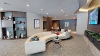 This single-floor space presents a refined, contemporary lounge environment defined by thoughtful design and high-end finishes. The centerpiece of the main seating area is a sweeping curved white sectional sofa, elegantly arranged around a sculptural glass and metal coffee table adorned with a vibrant floral arrangement. Decorative teal accent pillows complement the neutral palette and add a subtle pop of color.

The flooring features a patterned carpet with an organic, flowing motif that anchors the seating area and adds visual texture to the space. Recessed lighting throughout the ceiling provides warm, even illumination, enhancing the room's polished ambiance. A warm wood-paneled column serves as an architectural focal point, connecting the primary lounge area to a secondary seating zone visible in the background, which features darker accent walls and additional intimate furniture groupings.

Built-in shelving along one wall displays curated décor items, books, and sculptures, contributing to the space's cultivated character. Framed contemporary artwork is positioned throughout, reinforcing the sophisticated, gallery-like atmosphere. The overall design balances openness with intimate conversation areas, creating a welcoming and upscale communal environment.