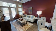 This well-appointed single-floor residence makes a striking first impression with its thoughtfully designed living area. The space is anchored by a dramatic deep crimson accent wall that adds depth and visual contrast against the neutral, warm-toned walls throughout. Large windows allow generous amounts of natural light to pour in, brightening the light wood-style flooring that runs underfoot and enhancing the overall sense of openness.

The living area is elegantly furnished with a tufted cream sofa, complementary accent chairs in rust and white tones, and a geometric-patterned area rug in warm oranges and golds that ties the room together. A sleek, industrial-style coffee table serves as a functional centerpiece, while matching black-and-white chevron table lamps on either side of the sofa provide stylish ambient lighting. A framed piece of artwork above the accent wall adds a curated, finished touch.

The ceiling features a ceiling fan, offering both practical airflow and a subtle design element. A sliding glass door or large window near the seating area hints at outdoor access, further connecting the interior to the outside environment. The overall ambiance is modern yet cozy, blending bold design choices with comfortable, livable furnishings.