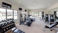 This single-level fitness center offers a spacious, open layout designed to accommodate a wide range of exercise needs. The room features neutral-toned carpet flooring, white walls, and recessed lighting that creates a clean, bright environment conducive to focused workouts.

Large windows along multiple walls allow generous amounts of natural light to fill the space while providing outdoor views, contributing to an open and energizing atmosphere. Wall-mounted televisions are positioned for easy viewing from cardio equipment.

The cardio zone is well-stocked with multiple treadmills and elliptical trainers arranged in orderly rows. Strength training options are equally comprehensive, including cable resistance machines, a functional trainer, weight benches, a dumbbell rack, and various selectorized machines for targeted muscle groups. A water cooler station is conveniently located within the room.

Large mirrors are mounted along one wall, visually expanding the space and aiding users in monitoring their form. A dedicated free weights and stretching area provides additional flexibility for varied workout routines. The fitness center is accessed via a glass-paneled door, and safety features including fire extinguishers and clearly marked exits are present throughout.