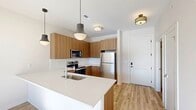 This single-floor unit showcases a clean, modern design throughout, with light wood-tone flooring connecting the main living areas and crisp white walls creating a bright, airy atmosphere. The kitchen is a standout feature, equipped with flat-front wood-grain cabinetry in a warm walnut finish, stainless steel appliances including a refrigerator, electric range, and over-the-range microwave, and a white quartz peninsula with an integrated sink and pendant lighting overhead. The layout flows naturally into the living area, which receives abundant natural light through large sliding glass doors that open onto a private balcony with views of the surrounding neighborhood.

The bedrooms are carpeted for comfort, with one featuring a ceiling fan, generous windows, and direct balcony access. A walk-in closet with wrap-around hanging rods provides ample storage. The bathroom includes wood-look tile flooring and a spacious walk-in shower with sliding glass doors and chrome fixtures. A dedicated laundry area houses a stacked Whirlpool washer and dryer alongside a water heater and electrical panel. Throughout the unit, the design balances functionality with contemporary aesthetics.
