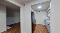 This single-story unit offers a well-organized layout with distinct living, kitchen, bedroom, and bathroom spaces connected by consistent design elements throughout. The living room serves as a central gathering area, highlighted by a white-surround fireplace and rich, dark wood-style flooring that extends through much of the unit. A textured ceiling adds dimension overhead, while natural light enters through windows fitted with horizontal blinds.

The kitchen is efficiently arranged with grey cabinetry, granite-look laminate countertops, and stainless-style appliances including a dishwasher and black refrigerator. Wood-style flooring continues here, maintaining visual continuity from the living areas. A pass-through layout connects the kitchen to the adjacent hallway and rooms.

The bedroom features soft carpeting, a ceiling fan with integrated lighting, and a window that allows in natural light, creating a comfortable and functional space. An additional room off the main living area offers flexibility as a secondary bedroom or office.

The bathroom includes a grey vanity with a wide mirror, Hollywood-style light bar, and speckled countertop surface, complemented by neutral carpet and access to a separate shower and toilet area. Throughout the unit, neutral grey-toned walls, white trim, and consistent flooring choices create a clean, cohesive aesthetic.