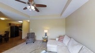 This single-floor residence offers a thoughtfully designed living space with a welcoming and comfortable ambiance. The living room serves as the centerpiece of the home, highlighted by a distinctive tray ceiling that adds architectural depth and elegance to the room. A ceiling fan with a light fixture is centrally mounted, providing both function and style. The walls are finished in soft, neutral tones that create a calm and inviting atmosphere. Plush carpeting underfoot adds warmth and comfort to the space. The room comfortably accommodates a sectional sofa, an accent armchair with a patterned upholstery, and a glass-top side table, reflecting a cozy and livable layout. A table lamp provides warm ambient lighting, complementing the overall tone of the interior. A partial view into an adjacent area suggests a connected kitchen or hallway, hinting at a practical, open flow between rooms. The combination of classic architectural details, neutral finishes, and comfortable furnishings gives this residence a relaxed yet refined character, making it a pleasant and functional place to call home.