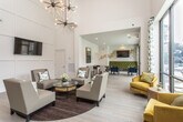 Located in the prestigious West Paces Ferry neighborhood of Atlanta, Georgia, this single-floor community clubhouse delivers an elevated residential amenity experience through thoughtful design and high-end finishes. The space is anchored by a dramatic great room with soaring double-height ceilings, paneled white walls, and light hardwood flooring that runs throughout, creating a bright and airy atmosphere. A striking sputnik chandelier serves as a centerpiece overhead, complementing the modern aesthetic. The lounge area features plush neutral seating arrangements and gold accent chairs positioned around a glass-top coffee table, ideal for relaxed gatherings. Floor-to-ceiling windows framed by bold patterned drapes flood the interior with natural light and offer direct views of the outdoor swimming pool and surrounding courtyard. A central bar and kitchen area showcases a long white waterfall-edge island with bar seating, rich dark wood cabinetry, a built-in sink, and a dramatic marble slab accent wall with a mounted flat-screen television. An adjacent game and dining zone features tufted green banquette seating, bar-height tables, and decorative wall art, providing a versatile entertainment space. Together, these areas create a cohesive, sophisticated clubhouse environment designed for both social interaction and quiet relaxation.