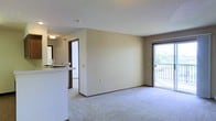 This single-floor unit offers a practical and comfortable open-concept layout with a clean, neutral aesthetic. The main living area is finished with soft wall-to-wall carpeting and painted in light, neutral tones that lend a bright and welcoming ambiance. A sliding glass door provides direct access to a private balcony, allowing natural light to filter into the space and offering views of the surrounding greenery.

Adjacent to the living area, a kitchen is visible behind a raised breakfast bar, which serves as a natural divider between the two spaces. The bar provides additional seating and counter space, making it a functional element for casual dining or entertaining. Glimpses of glassware and countertop details suggest a well-equipped kitchen setup.

A doorway leading to additional rooms of the unit is visible from the main living space, hinting at further accommodations beyond the open area. Textured ceilings are consistent throughout, and dark-trimmed door frames provide subtle contrast against the light walls. Overall, the space presents a straightforward, functional interior well-suited for everyday living.