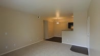 This single-floor residence offers a practical and open layout designed for comfortable everyday living. The living area is finished with plush wall-to-wall carpeting in a neutral tone, creating a warm and inviting atmosphere that flows seamlessly into the dining and kitchen spaces.

The transition from the living area to the dining space is marked by a shift to darker flooring, providing a subtle visual distinction between the two zones. A pendant light fixture hangs above the dining area, adding a touch of understated elegance and providing focused overhead lighting.

The kitchen is positioned adjacent to the dining area and features dark upper cabinetry paired with a raised breakfast bar counter, offering both additional prep space and a casual seating option. A stainless steel appliance is visible within the kitchen, complementing the darker cabinetry tones.

Throughout the space, the walls are finished in a soft, warm beige, contributing to a cohesive and neutral color palette. Visible HVAC vents and clean white baseboards speak to the functional infrastructure of the unit. The overall ambiance is understated and move-in ready, providing a versatile blank canvas well-suited for a range of interior styles and furnishing preferences.