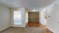 This single-floor space offers a welcoming interior defined by a thoughtful mix of materials and textures. The main living area transitions between warm terracotta tile flooring and plush carpeting, creating distinct zones within an open layout. A striking exposed brick accent wall serves as a standout architectural feature, adding character and a rustic warmth to the space. Multiple windows fitted with horizontal blinds allow for generous natural light while maintaining privacy, giving the interior a bright and airy feel. The textured popcorn ceiling adds a classic finish overhead, while a flush-mount ceiling light provides functional illumination. A doorway visible from the main area leads to an adjacent room with matching brick detailing, suggesting a consistent design language throughout. To the side, a pass-through or entryway hints at a connected kitchen or utility space, also featuring brick tile elements that tie the overall aesthetic together. Neutral wall tones complement the earthy flooring and brick, creating a cohesive, grounded ambiance. The layout flows naturally between spaces, making the interior feel connected and versatile for a variety of uses.