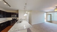 This single-floor residence offers a thoughtfully designed open-concept layout that seamlessly connects the kitchen and living areas. The kitchen is a standout feature, showcasing rich dark cabinetry with ample upper and lower storage, granite-look countertops, and a full suite of stainless steel appliances including a gas range, microwave, and refrigerator. A peninsula with a double sink and elegant pendant lighting serves as both a functional workspace and a natural dividing point between the kitchen and the adjoining living space. The living area is generously proportioned, finished with plush wall-to-wall carpeting, a ceiling fan with integrated lighting, and a glass door that invites natural light from outside. The bedrooms continue the warm, neutral palette, each featuring carpet flooring, ceiling fans, and windows dressed with blinds. At least one bedroom includes a walk-in closet for convenient storage. The bathrooms are finished with dark cabinetry consistent with the kitchen, white countertops, and hardwood-style flooring. The primary bathroom features a dual-sink vanity with a large mirror and well-appointed overhead lighting. Throughout, the space maintains a cohesive, modern aesthetic with neutral tones, clean lines, and practical finishes.