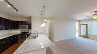 This single-floor residence offers a thoughtfully designed open-concept layout that seamlessly connects the kitchen and living areas. The kitchen is a standout feature, showcasing rich dark cabinetry with ample upper and lower storage, granite-look countertops, and a full suite of stainless steel appliances including a gas range, microwave, and refrigerator. A peninsula with a double sink and elegant pendant lighting serves as both a functional workspace and a natural dividing point between the kitchen and the adjoining living space. The living area is generously proportioned, finished with plush wall-to-wall carpeting, a ceiling fan with integrated lighting, and a glass door that invites natural light from outside. The bedrooms continue the warm, neutral palette, each featuring carpet flooring, ceiling fans, and windows dressed with blinds. At least one bedroom includes a walk-in closet for convenient storage. The bathrooms are finished with dark cabinetry consistent with the kitchen, white countertops, and hardwood-style flooring. The primary bathroom features a dual-sink vanity with a large mirror and well-appointed overhead lighting. Throughout, the space maintains a cohesive, modern aesthetic with neutral tones, clean lines, and practical finishes.