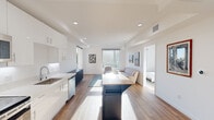 This thoughtfully designed single-floor unit offers a clean, contemporary aesthetic throughout its six distinct spaces. The open-concept layout connects a well-appointed kitchen to a bright living area, creating a seamless flow ideal for everyday living. The kitchen features floor-to-ceiling white cabinetry, stainless steel appliances including a refrigerator, range, microwave, and dishwasher, a deep undermount sink, and a butcher block-topped island with navy cabinetry. Light wood-look flooring runs continuously through the main living areas, complementing the all-white walls and recessed lighting overhead. The living room is bathed in natural light from large windows that frame lush greenery, lending a calm, airy ambiance to the space. Two well-proportioned bedrooms offer restful retreats, each featuring the same light hardwood flooring, crisp white walls, and generous windows with outdoor views. The full bathroom is finished with large-format marble-look wall tile surrounding the soaking tub and shower, a modern wood-grain vanity with white countertop, and warm wood flooring. A dedicated storage room with distinctive wood-look tile covering both the floor and walls rounds out the unit, offering practical functionality alongside refined design details throughout.