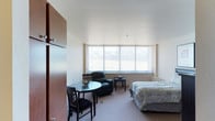 This single-floor studio unit is thoughtfully organized to maximize functionality within a compact footprint. The main living area serves as the central hub of the space, combining sleeping, lounging, and dining functions seamlessly. Carpeted flooring runs throughout, lending warmth underfoot, while white walls and a smooth ceiling create a clean, neutral backdrop. A large window fitted with vertical blinds spans much of the exterior wall, drawing in generous natural light and offering open outdoor views, contributing to an airy atmosphere despite the unit's modest scale. The sleeping area accommodates a twin bed positioned beside a nightstand and lamp, while a round dining table with chairs and a leather recliner provide comfortable living and dining options. A tall wardrobe cabinet and a dresser offer practical storage solutions. The galley-style kitchen is efficiently arranged with dark wood cabinetry above and below the counter, a stainless steel sink, a dishwasher, a microwave, and a full-size refrigerator, with additional counter space serving as a workspace or breakfast bar. The bathroom is straightforward in design, featuring a tub-shower combination with a curtain, a toilet, and a vanity with cabinetry below.