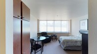 This single-floor studio unit is thoughtfully organized to maximize functionality within a compact footprint. The main living area serves as the central hub of the space, combining sleeping, lounging, and dining functions seamlessly. Carpeted flooring runs throughout, lending warmth underfoot, while white walls and a smooth ceiling create a clean, neutral backdrop. A large window fitted with vertical blinds spans much of the exterior wall, drawing in generous natural light and offering open outdoor views, contributing to an airy atmosphere despite the unit's modest scale. The sleeping area accommodates a twin bed positioned beside a nightstand and lamp, while a round dining table with chairs and a leather recliner provide comfortable living and dining options. A tall wardrobe cabinet and a dresser offer practical storage solutions. The galley-style kitchen is efficiently arranged with dark wood cabinetry above and below the counter, a stainless steel sink, a dishwasher, a microwave, and a full-size refrigerator, with additional counter space serving as a workspace or breakfast bar. The bathroom is straightforward in design, featuring a tub-shower combination with a curtain, a toilet, and a vanity with cabinetry below.
