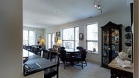 This single-floor space offers an open and well-connected layout that seamlessly blends dining and living areas into one cohesive environment. Large windows line the walls, flooding the interior with natural light and framing views of the surrounding urban cityscape, creating a bright and airy atmosphere throughout.

The dining area is anchored by a substantial dark wood table paired with matching high-back chairs, lending a classic and formal character to the space. Adjacent to it, a circular table with cushioned armchairs creates a more intimate sitting or game area, ideal for casual gatherings. The warm, neutral-toned carpet softens the overall aesthetic and adds comfort underfoot.

A tall, dark wood display cabinet stands prominently against one wall, showcasing a curated collection of decorative objects and glassware, adding a personal and elegant touch. Framed artwork adorns the walls, contributing to the refined and lived-in character of the space. Track lighting on the ceiling supplements the abundant natural light, ensuring the interior remains well-lit at all times.

The clean white walls and smooth ceilings provide a neutral backdrop that allows the rich, dark furnishings to stand out. Overall, the space exudes a classic, welcoming ambiance with thoughtful attention to detail and a comfortable, functional arrangement suited for both entertaining and everyday living.