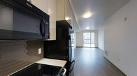 This modern single-floor residence showcases a thoughtfully designed open-concept layout that seamlessly connects the kitchen and living areas. The kitchen is outfitted with black appliances, including a refrigerator, electric range, and over-the-range microwave, complemented by cream high-gloss cabinetry that extends to the ceiling for ample storage. A decorative tile backsplash in muted earth tones adds texture and visual interest to the cooking space, while light-colored countertops provide a clean contrast. The flooring throughout is a smooth, grey wood-look surface that flows consistently from the kitchen into the open living area, lending a cohesive and contemporary feel to the space. The living area is bright and airy, bathed in natural light streaming through a large floor-to-ceiling window that frames views of the surrounding urban environment. Recessed flush-mount ceiling lights provide even ambient illumination. The crisp white walls and minimalist architectural details create a neutral, versatile backdrop well-suited for a variety of interior styles. The overall ambiance is clean, modern, and functional, reflecting a well-appointed urban living space designed with both style and practicality in mind.