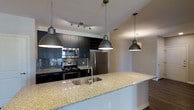 This single-floor space showcases a thoughtfully designed, open-concept kitchen that serves as the heart of the layout. The kitchen is outfitted with a suite of stainless steel appliances, including a gas range, built-in microwave, and a full-sized refrigerator, all set against striking dark cabinetry that creates a bold, modern contrast. A textured mosaic tile backsplash adds visual depth and a refined aesthetic to the cooking area, complemented by a dedicated track lighting system overhead. The expansive island takes center stage, featuring a light-toned granite countertop with natural veining and an undermount stainless steel sink — providing ample workspace and a natural gathering point. Three matching industrial-style dome pendant lights hang above the island, reinforcing the contemporary design language throughout. The open layout flows seamlessly into the adjacent living area, creating a connected, airy feel. Neutral wall tones and clean lines keep the atmosphere polished and inviting, making this a highly functional yet visually refined space well-suited for both everyday living and entertaining.