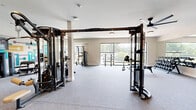 Located at 1351 East Woodlawn Road in Charlotte, NC, this fitness center offers a generously sized, single-floor exercise space designed for a comprehensive workout experience. The open floor plan provides ample room to move freely between stations, with neutral-toned carpet flooring throughout that balances comfort and functionality.

The space is outfitted with an impressive array of equipment, including a multi-station cable machine system, a power rack, a full dumbbell rack with flat benches, and multiple selectorized strength machines for targeted muscle training. Ceiling fans are mounted throughout to maintain airflow, and flush-mount and pendant lighting ensure the space remains well-lit.

Large windows along the perimeter fill the room with natural light and offer views of the surrounding greenery, contributing to an open and energizing atmosphere. Full-length mirrors are positioned along one wall, enhancing the sense of space while supporting proper form during workouts. Mounted flat-screen TVs provide entertainment throughout training sessions.

A standout design feature is the vibrant geometric mural in teal, yellow, burgundy, and white adorning one wall, adding a bold artistic focal point to the otherwise clean, modern aesthetic. Patterned glass partition doors with decorative frosted panels offer a stylish visual divide, contributing to the polished, contemporary feel of this well-appointed fitness facility.