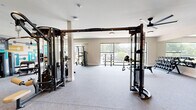 Located at 1351 E Woodlawn Rd in Charlotte, NC, this well-appointed fitness center offers a generous, open floor plan designed to accommodate a comprehensive workout experience. The space is thoughtfully organized across a single level, providing ample room for both strength training and functional movement.

The interior features a combination of carpet and rubberized flooring, ceiling fans, and flush-mounted lighting that create a comfortable and practical environment. Large windows along the perimeter flood the space with natural light and offer views of the surrounding greenery, enhancing the overall atmosphere.

A standout feature is the full-length mirrored wall, which amplifies the sense of space and supports proper form during exercise. The strength training area is equipped with multi-functional cable machines, a comprehensive dumbbell rack with flat benches, and a variety of selectorized weight machines positioned throughout the room. A large flat-screen television is mounted for entertainment during workouts.

Distinctive decorative elements include a bold, geometric mural in teal, yellow, and purple tones, as well as stylized patterned glass partitions along one wall, adding a modern and vibrant aesthetic to the space. The open layout and diverse equipment selection make this fitness center a well-rounded facility for residents and users of all fitness levels.