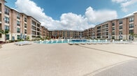 Located at 1724 Partnership Way in Houston, TX, this amenity space is part of a contemporary multi-story residential complex that blends brick, stone, and modern architectural detailing to create an upscale resort-like atmosphere. The living area opens to a sweeping outdoor courtyard finished in smooth, neutral-toned concrete that serves as the heart of the community.

The centerpiece of the outdoor space is a generously sized resort-style swimming pool with a shallow sun shelf fitted with sleek white lounge chairs, allowing residents to relax both in and out of the water. The pool deck surrounds the water with ample space for additional chaise lounges and umbrella-shaded seating.

A covered outdoor kitchen and dining pavilion provides an ideal setting for gatherings, featuring stainless steel grills, a full preparation counter, and a dining table with modern white chairs beneath a vaulted ceiling with ceiling fan. The warm brick and stone accent walls lend a refined, yet inviting character to the space.

Lush landscaping throughout the courtyard includes palm trees, colorful ornamental plantings, decorative rock borders, and manicured green lawns with lounge seating, creating a vibrant, tropical ambiance. The multi-story building facades frame the courtyard with private balconies, large windows, and clean architectural lines that enhance the sense of openness and community throughout this well-appointed space.