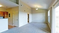 This single-floor residence offers a well-designed layout with a comfortable and functional living environment. The main living area is defined by soaring vaulted ceilings that create a sense of openness and airiness, complemented by neutral-toned walls that lend a clean, versatile aesthetic throughout. Soft, plush wall-to-wall carpeting covers the floors, adding warmth and comfort underfoot. A pendant light fixture anchors the space with practical overhead illumination, while HVAC vents integrated into both the walls and ceiling ensure year-round climate comfort. The open layout flows naturally toward adjacent spaces, with interior doors providing access to what appears to be a utility or secondary room and a connecting hallway leading further into the residence. White six-panel doors with classic hardware add a traditional architectural touch. The combination of vaulted ceilings, neutral finishes, and a spacious footprint gives the main living area a bright and inviting ambiance, making it well-suited for a variety of furniture arrangements and living styles. The overall design prioritizes simplicity and practicality, offering a move-in-ready canvas for personalization.
