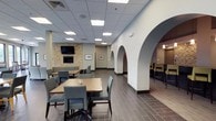 This community room offers a welcoming and well-appointed gathering space designed for shared use. The room's most distinctive architectural feature is a series of curved white arches that elegantly divide the space into distinct zones, creating a sense of depth and visual interest while maintaining an open feel throughout.

The main dining area is furnished with wooden tables and upholstered gray chairs arranged to encourage conversation and group dining. A stone-accented accent wall serves as a focal point, complemented by a wall-mounted flat-screen television, adding both style and functionality to the lounge area.

Toward the rear, a separate seating zone—accessible through the arched openings—features counter-height tables and bar stools, providing an alternative setting for more casual interaction. The flooring throughout consists of large-format dark tile, lending a clean and modern aesthetic that ties the space together.

Recessed panel lighting in the drop ceiling provides consistent, even illumination across all areas. Framed artwork on the walls adds a refined touch, contributing to an overall ambiance that is both practical and comfortable for residents and guests.