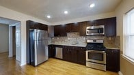 This single-floor space showcases a thoughtfully designed kitchen that combines functionality with modern aesthetics. The kitchen is anchored by a full suite of stainless steel appliances, including a full-size refrigerator, a gas range with oven, a built-in microwave, and a dishwasher — offering a complete and practical cooking environment.

Dark espresso-toned cabinetry lines both the upper and lower walls, providing generous storage capacity. The cabinetry features clean, shaker-style door fronts with brushed metal hardware, lending a contemporary look. Light-toned granite or stone countertops contrast beautifully against the dark cabinets, and the neutral stone tile backsplash adds visual texture while complementing the overall color palette.

The sink is centrally positioned along the counter run, beneath recessed lighting that illuminates the workspace effectively. Light hardwood flooring extends throughout, adding warmth and continuity to the space. The L-shaped layout of the kitchen maximizes counter and cabinet space while maintaining an open and navigable flow. Recessed ceiling lights provide consistent, practical illumination throughout the area.