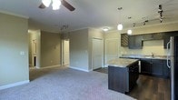 This single-story space showcases an open-concept layout that seamlessly connects the living area and kitchen. The living room features neutral-toned carpeting and is anchored by a ceiling fan with light fixture, creating a comfortable and well-lit environment. Crown molding runs along the upper perimeter, adding a refined architectural detail to the space.

The kitchen is outfitted with dark espresso cabinetry that provides a bold contrast against the light-colored walls. A central island with a granite-style countertop offers additional workspace and casual seating potential. Pendant lighting hangs above the island, providing focused task lighting, while a decorative wine rack built into the upper cabinetry adds a distinctive design element.

The transition zone between the living and kitchen areas features hardwood-style flooring, distinguishing the two spaces while maintaining visual continuity. Multiple closet doors are visible along the back wall, offering generous storage capacity. The neutral warm-toned walls throughout contribute to a cohesive and inviting ambiance, while recessed and track lighting provide versatile illumination options across the space.