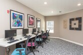 The Computer Center at Harmony at Conyers, located on Iris Drive SE in Conyers, Georgia, is a thoughtfully designed communal workspace configured to meet residents' technology needs. The single-floor space features a row of individual computer workstations lined along a sleek, light-colored desk surface, each equipped with a monitor, keyboard, and ergonomic black task chair. A window positioned near the workstation area allows natural light to filter into the room, contributing to a comfortable working environment.

A dedicated built-in credenza with rich dark wood cabinetry and a white countertop serves as a printing and document management station, housing a printer for resident use. Posted rules and guidelines for the Computer Center are prominently displayed near the entrance, ensuring clear communication of usage policies.

The space is finished with neutral-toned walls, patterned low-pile carpeting, and recessed lighting overhead. Decorative wall art, including bold floral framed prints and ornamental metallic wall sculptures, adds visual interest and warmth to the otherwise functional environment. An interior window provides a visual connection to an adjacent space. Overall, the Computer Center offers a clean, organized, and welcoming atmosphere suited for productivity.